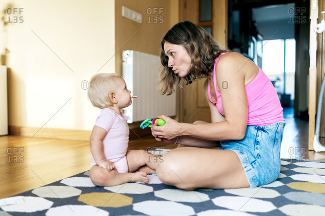 Side view of happy young woman in casual clothes playing with adorable little daughter while sitting on floor at home