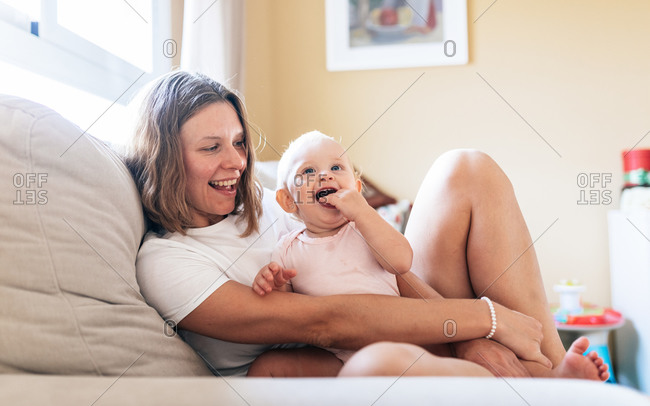 Happy adult woman hugging cute baby daughter sucking fingers while resting on soft couch at home