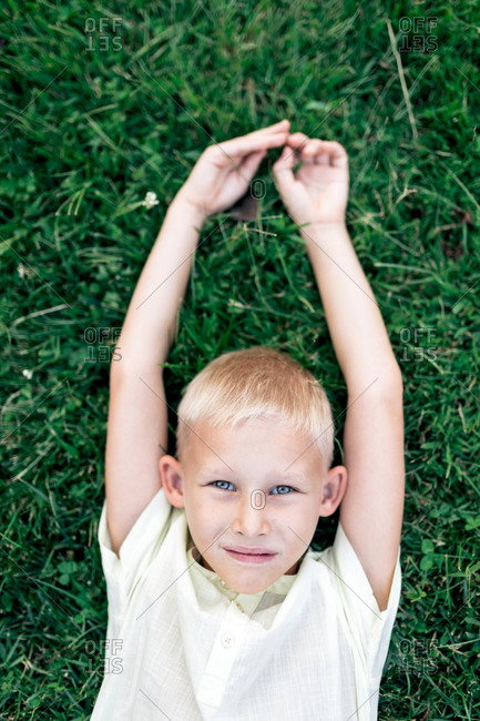 Overhead cheerful blond haired boy in white shirt looking at camera with smile while chilling on verdant grass with arms behind head