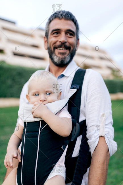 Cheerful adult man in casual clothing with beard looking ahead with toothy smile while carrying laughing infant with sling in park in daylight