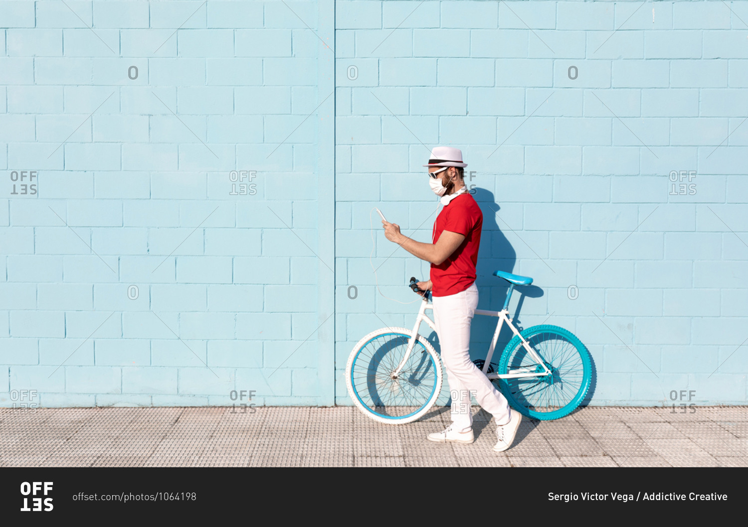 Side view of unrecognizable young guy in casual outfit and medical mask using mobile phone while standing near stylish bicycle and blue wall on city street