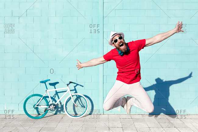Full body of expressive young bearded male hipster in trendy outfit and sunglasses jumping with outstretched arms and screaming near bicycle leaning on blue wall on street