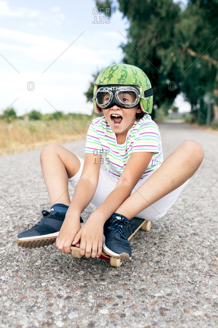 Ground level of content kid in watermelon helmet and goggles sitting on skateboard on asphalt roadway and looking away