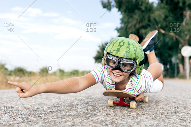 Ground level of happy kid in protective eyewear and ornamental watermelon helmet lying on skateboard on roadway and looking away