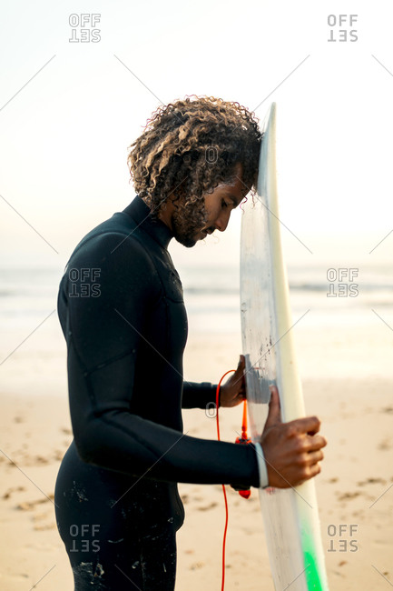 Side view of thoughtful ethnic male with curly hair in comfortable swimsuit holding surfboard at sunset