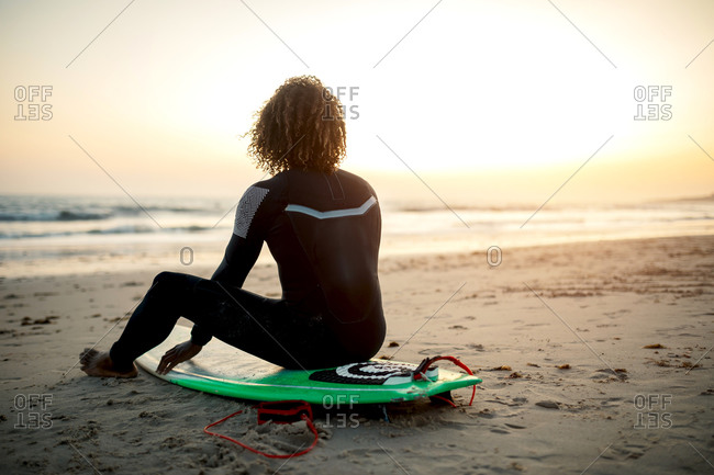 Full body back view of male surfer with fluffy hair in black wetsuit looking at sunset above ocean while resting on coast covered with sand