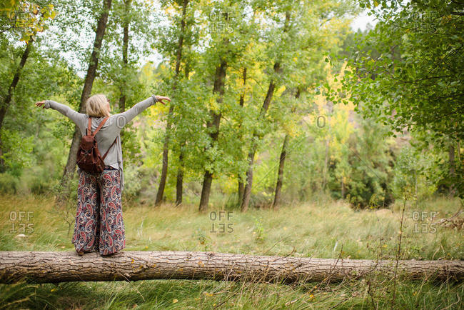 Full body of back view of unrecognizable content blonde female traveler in stylish outfit balancing on fallen tree trunk with hands apart while enjoying summer day in green forest