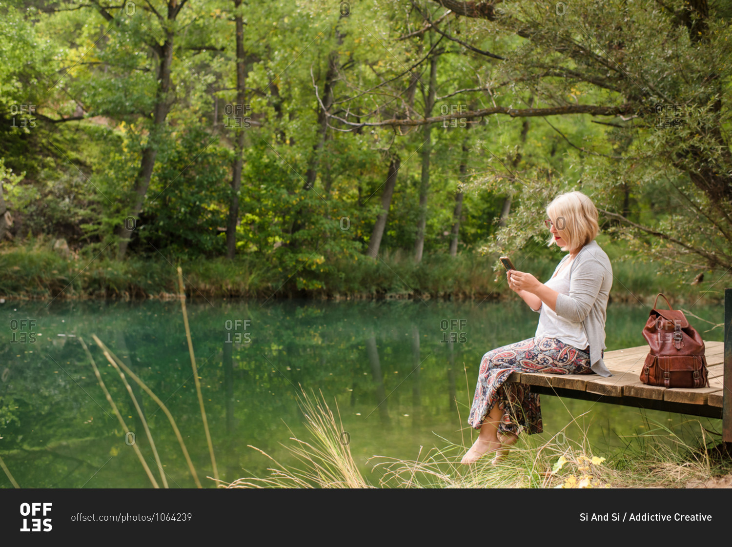 Side view of cheerful barefoot female in casual outfit sitting on wooden platform near calm pond and using smartphone while resting in green forest in summer day