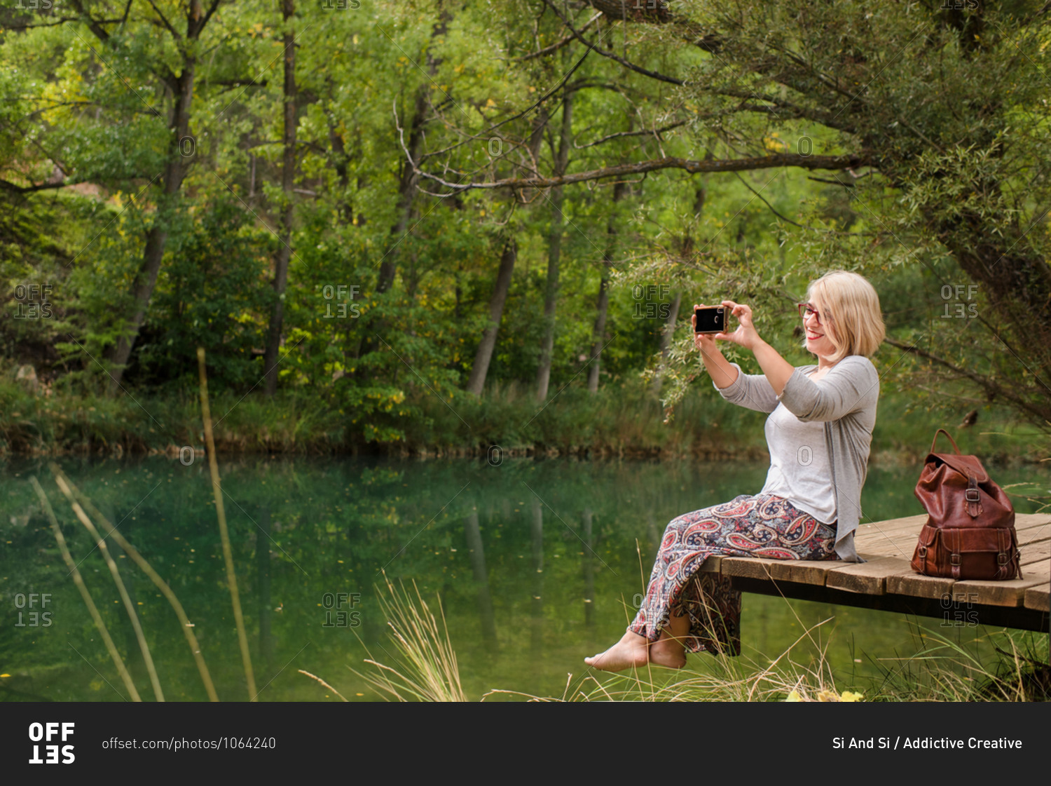 Side view of cheerful barefoot female in casual outfit sitting on wooden platform near calm pond and taking pictures of nature with smartphone while resting in green forest in summer day
