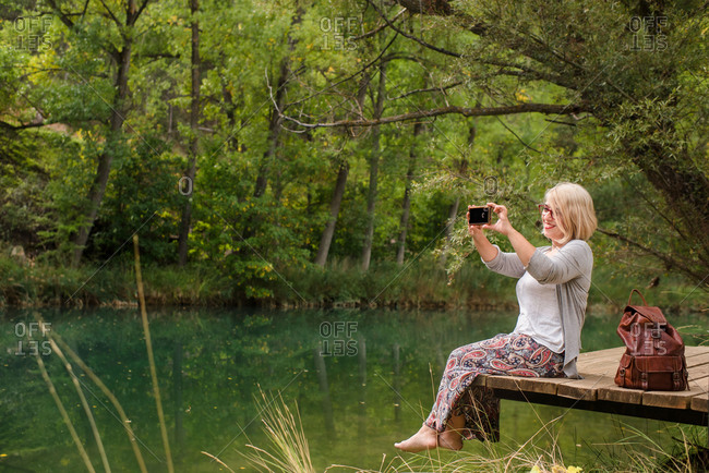 Side view of cheerful barefoot female in casual outfit sitting on wooden platform near calm pond and taking pictures of nature with smartphone while resting in green forest in summer day