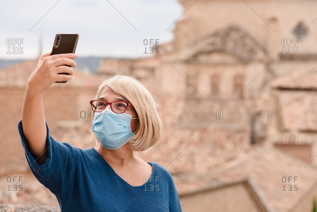 Adult female traveler in eyeglasses and medical mask for coronavirus prevention taking pictures of medieval architecture while standing against blurred exterior of old building in Cuenca town in Spain