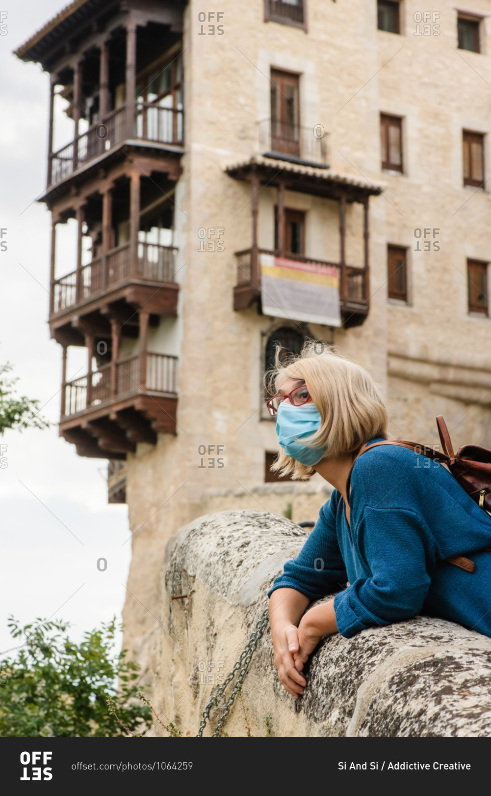 Low angle side view of travelling woman in protective mask leaning on stone border near old building and looking away while visiting medieval town Cuenca in Spain
