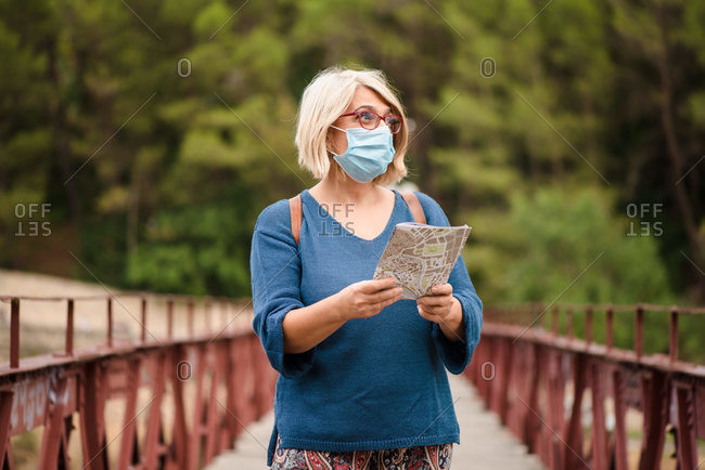 Adult female tourist holding map and looking away while standing on footbridge against green forest and searching for direction during travel in Spain