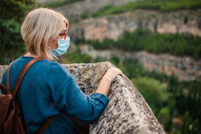 Side view of female tourist in medical mask standing near stone wall of medieval castle and admiring mountainous landscape while visiting Cuenca town in Spain