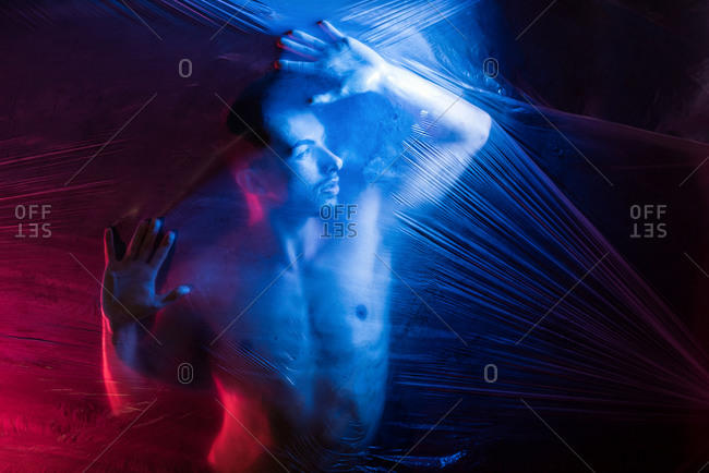 Pensive young handsome shirtless muscular male looking away while putting hands on surface of big transparent tape in light of blue and red lamps