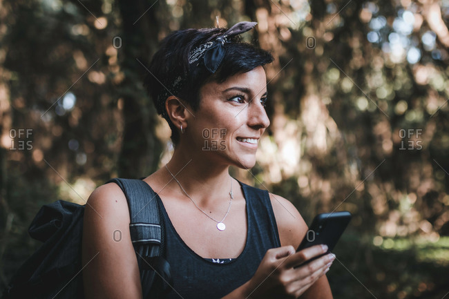 Portrait of a beautiful smiling woman with short hair and a beautiful smile holding her phone in the forest while hiking wearing a backpack in the woods looking away