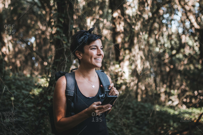 Portrait of a beautiful cheerful woman with short hair and a beautiful smile holding her phone in the forest while hiking wearing a backpack in the woods looking away