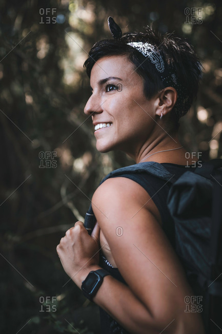 Close up portrait of a beautiful woman with short hair smiling in the forest while hiking and enjoying nature wearing a backpack in the woods looking away holding the strap