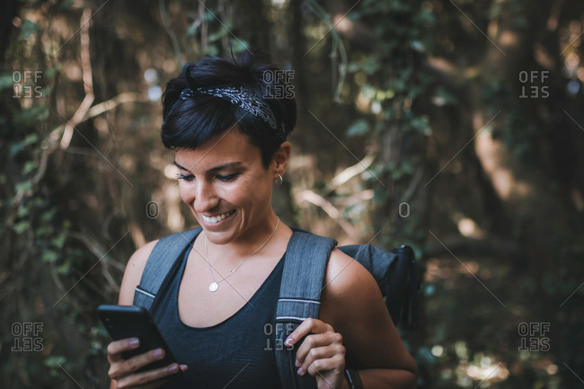 Portrait of a beautiful woman with short hair and a beautiful smile looking at her phone in the forest while hiking wearing a backpack