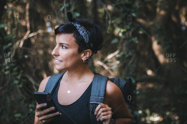 Portrait of a beautiful woman with short hair and a beautiful smile holding her phone looking away in the forest while hiking wearing a backpack