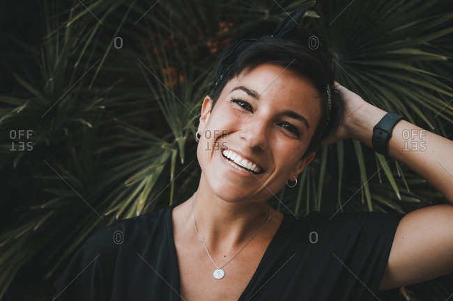 Portrait of a beautiful joyful woman with a beautiful smile and short hair looking at camera while smiling and touching her hair with plants in the background