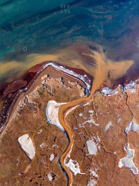 Aerial view of a yellow stream entering blue Olfusa river surrounded by frozen farmlands with red colored soil, Eyrarbakki, South Iceland