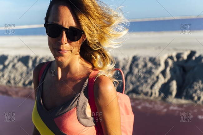 Pensive middle aged female traveler in sunglasses looking at camera while standing on blurred background of pink lake in Las Coloradas, Mexico