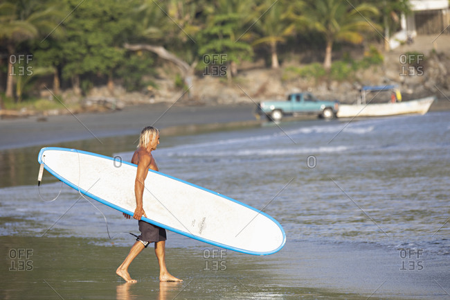 Sayulita, Nayarit, Mexico - June 18, 2018: Athletic senior man carrying surfboard on beach