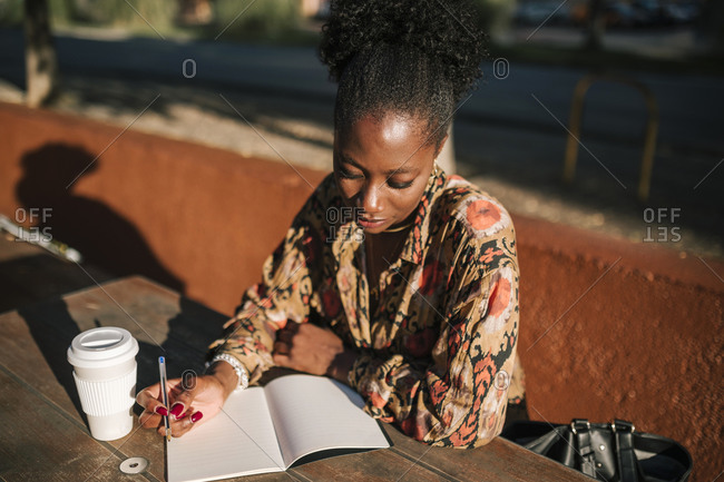 Young black woman writing notes in her agenda sitting in an outdoor cafe