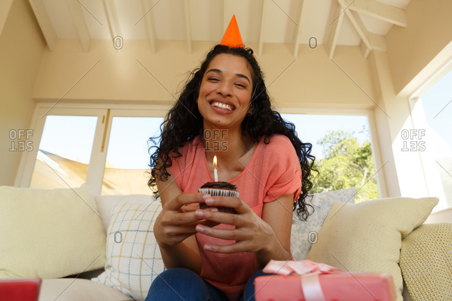 Mixed race woman celebrating birthday wearing party hat holding muffin with candle on it