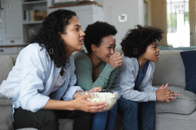 Mixed race lesbian couple and daughter sitting on couch watching tv and eating popcorn