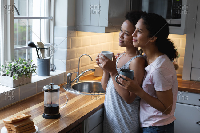 Smiling mixed race lesbian couple drinking coffee and embracing in kitchen
