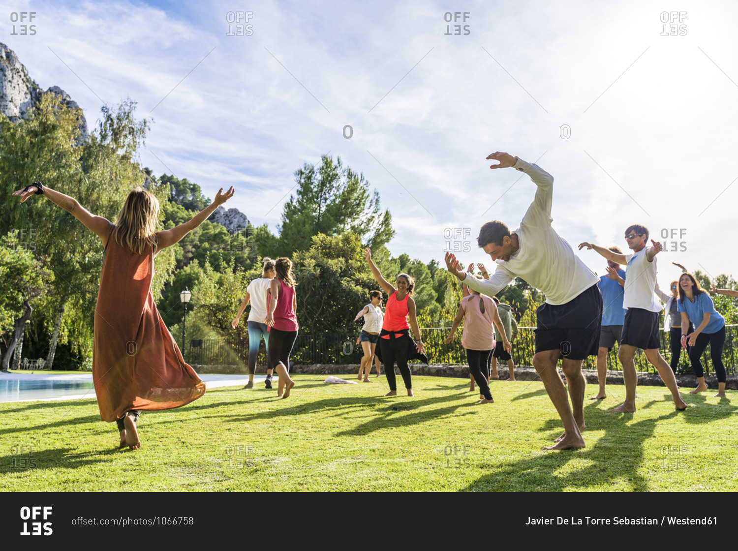 Mature female fitness instructor dancing with tourists at health retreat on grass against sky during sunny day