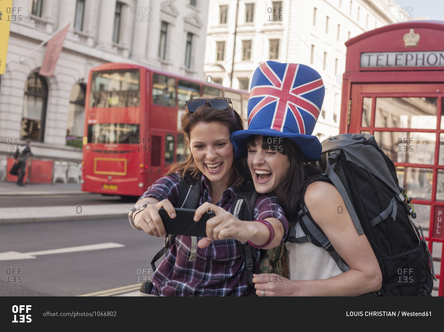 Happy woman taking selfie with friend wearing British flag hat against red telephone box in city