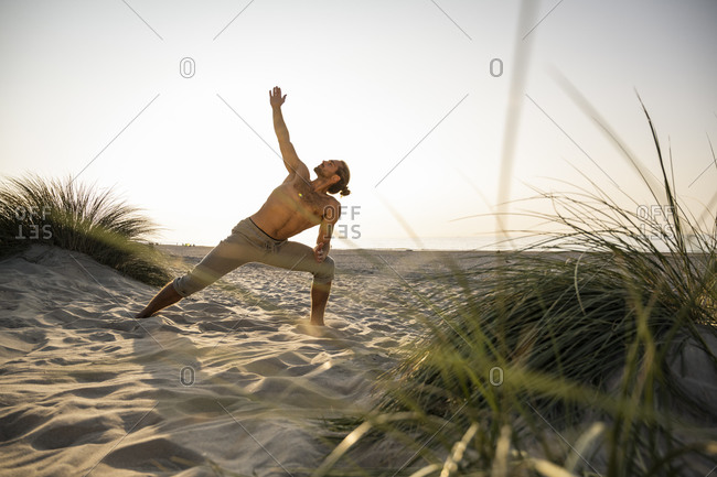 Shirtless young man practicing triangle position yoga at beach against clear sky during sunset
