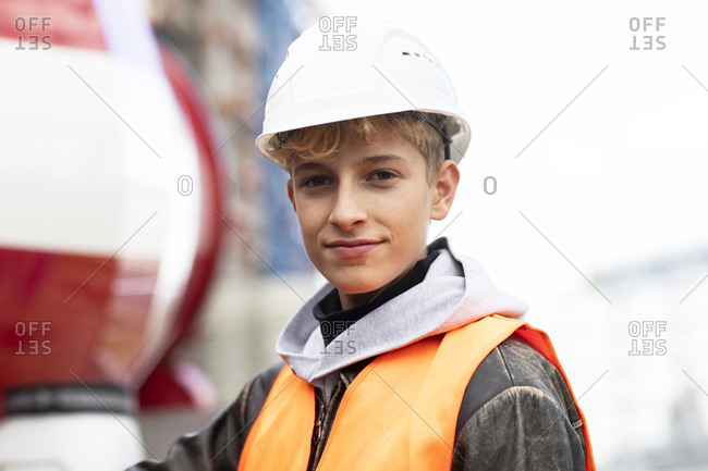 Confident male teenage trainee wearing hardhat and reflective clothing at construction site