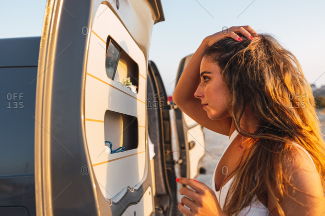 Young woman with hand in hair looking at mirror in camper van door while standing at beach