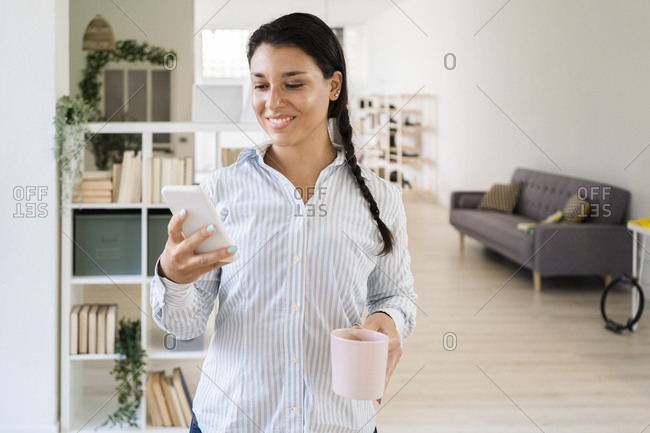 Smiling woman text messaging on smart phone while holding coffee cup standing at home