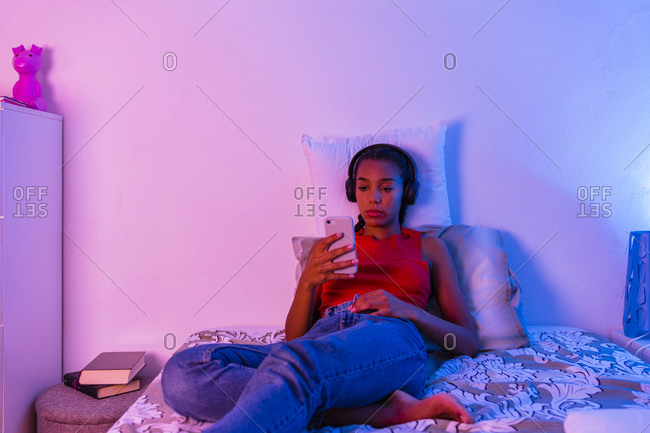 Teenage girl lying on bed using smart phone to listen music through headphone at home