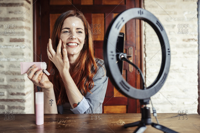 Smiling woman applying cream while filming make up tutorial on mobile phone at home