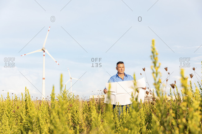 Male entrepreneur holding blueprint while standing against wind turbines on field