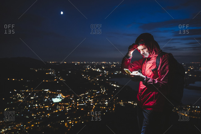 Mature male hiker using smart phone with illuminated city against sky at night