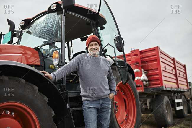 Smiling mature farmer standing with hand in pocket against red tractor at farm