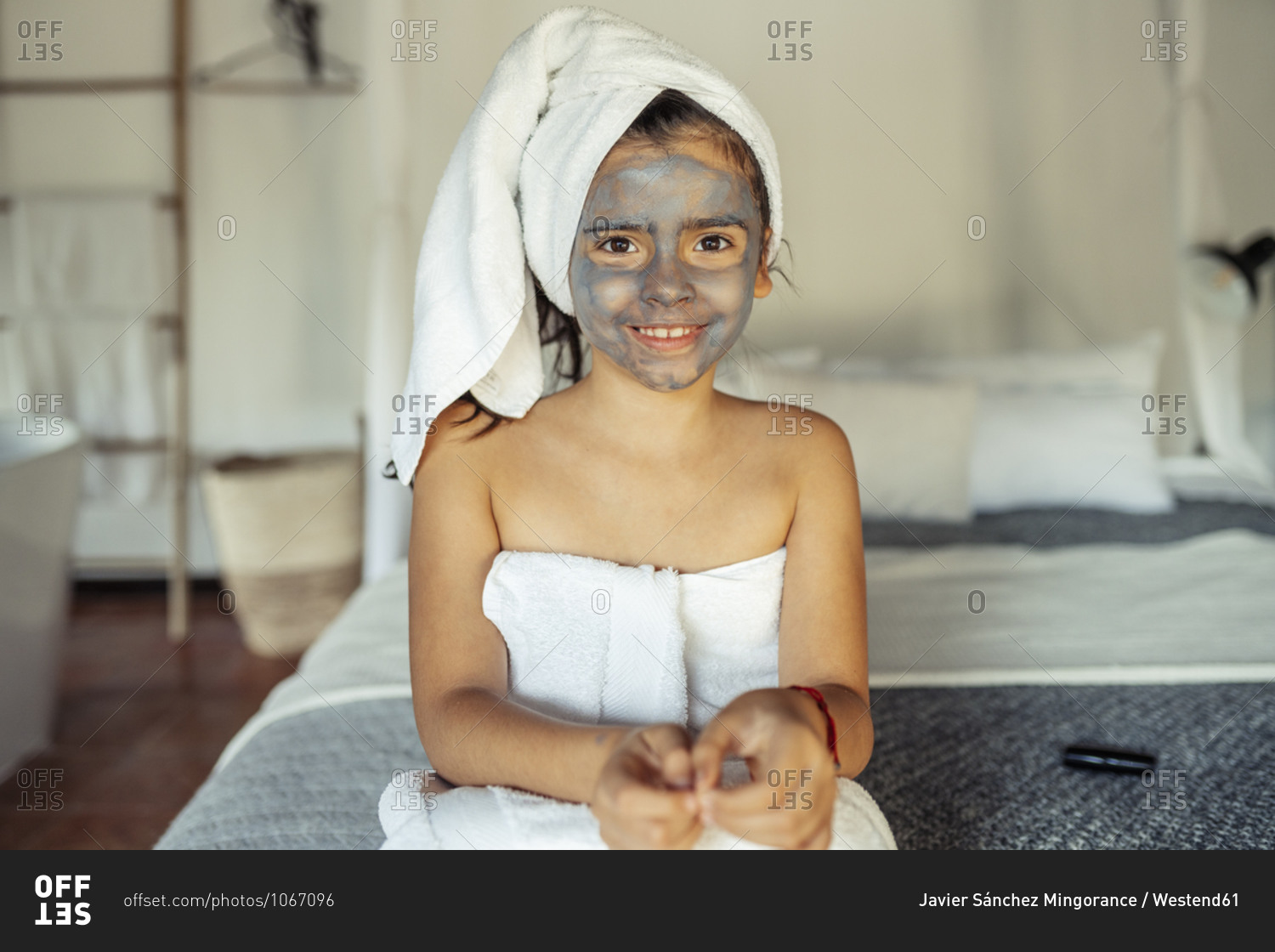 Smiling girl covered in towel with facial mask on face sitting on bed at home