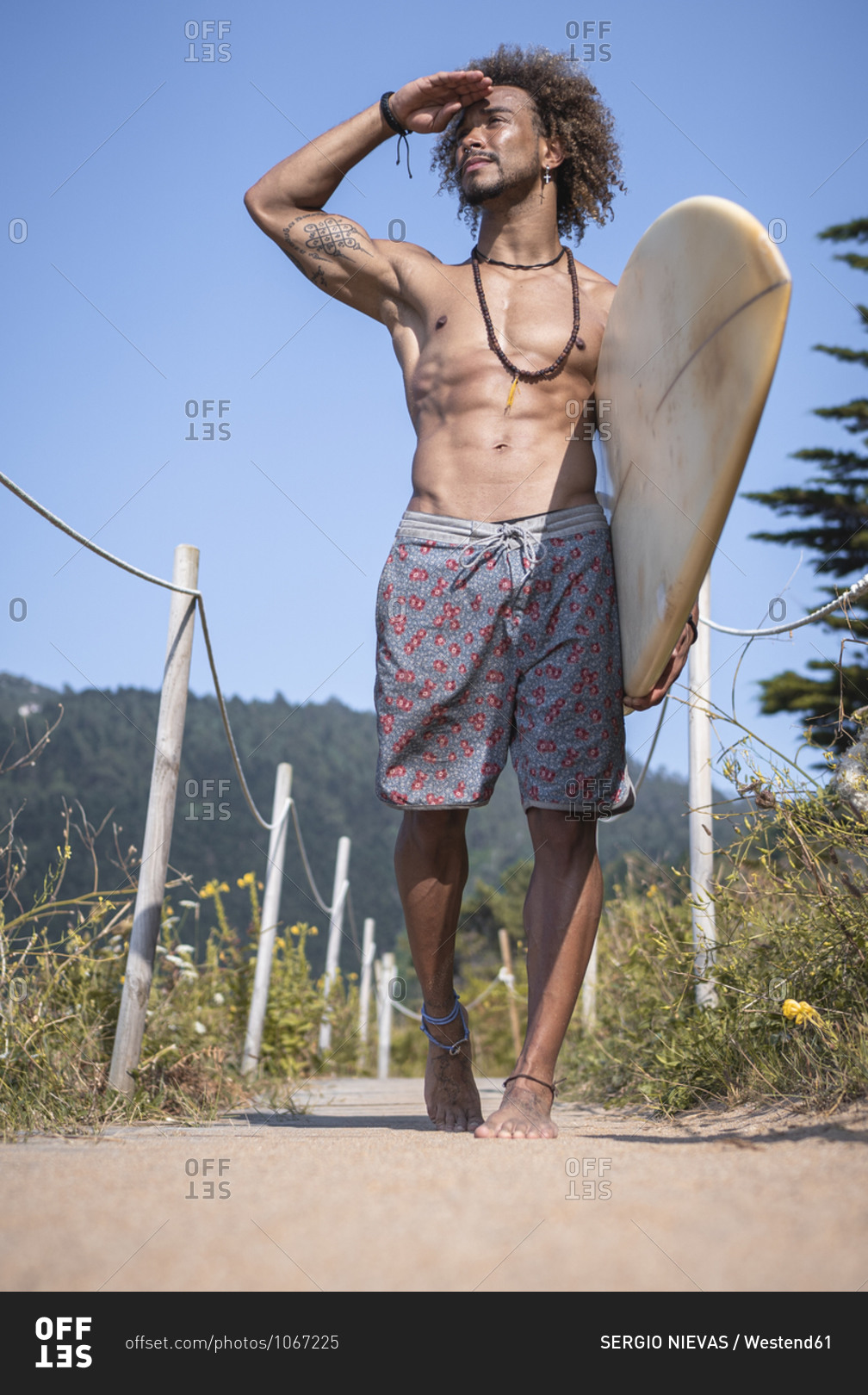 Shirtless young man with surfboard shielding eyes while walking at ...