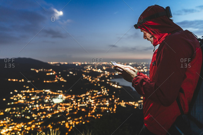 Male hiker using smart phone with illuminated city against sky at night