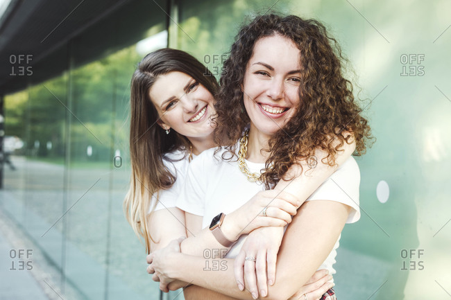 Smiling beautiful woman hugging female friend from behind against wall