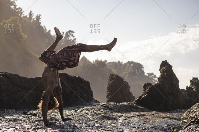 Young man practicing handstand on rock formation at beach against sky