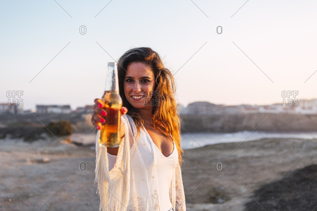 Smiling woman showing small white wine bottle at beach during sunset