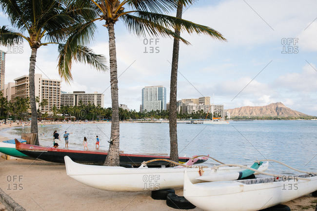 Honolulu, Oahu, Hawaii - March 17, 2020:  Boats and tourists on beach with palm trees and tall buildings in the background